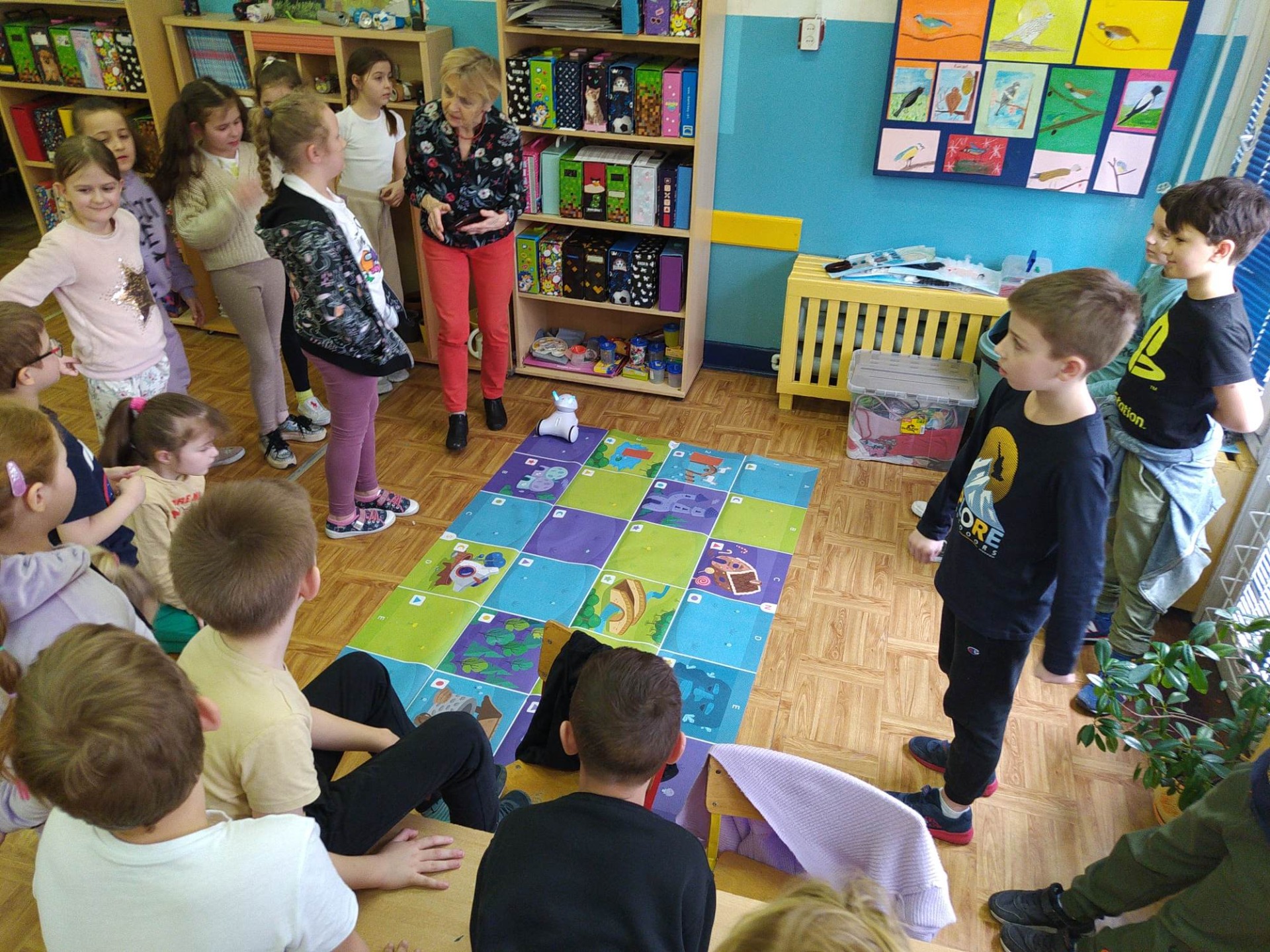 Dzieci zgromadzone wokół maty edukacyjnej. Programują robota Photon  i rozwiązują z nim zagadki literackie