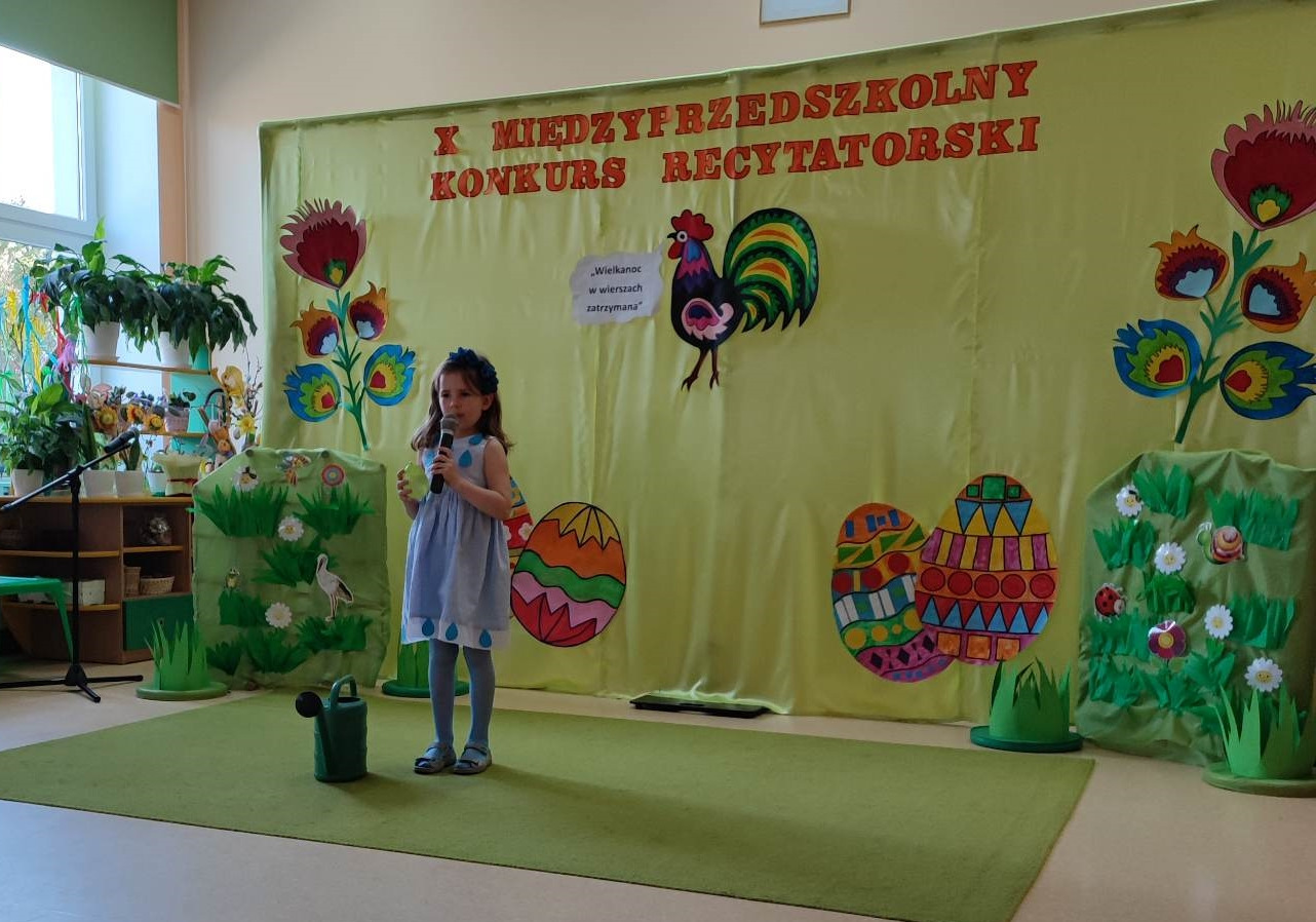 W srodkowej części fotografii stoi dziewczynka w wieku 6 lat stoi na zielonym dywanie z mikrofonem w ręku. Recytuje wiersz. W tle zielony materiał na którym znajdują się kolorowe pisanki i powieszony jest napis  X Międzyprzedszkolny Konkurs Recytatorski.
