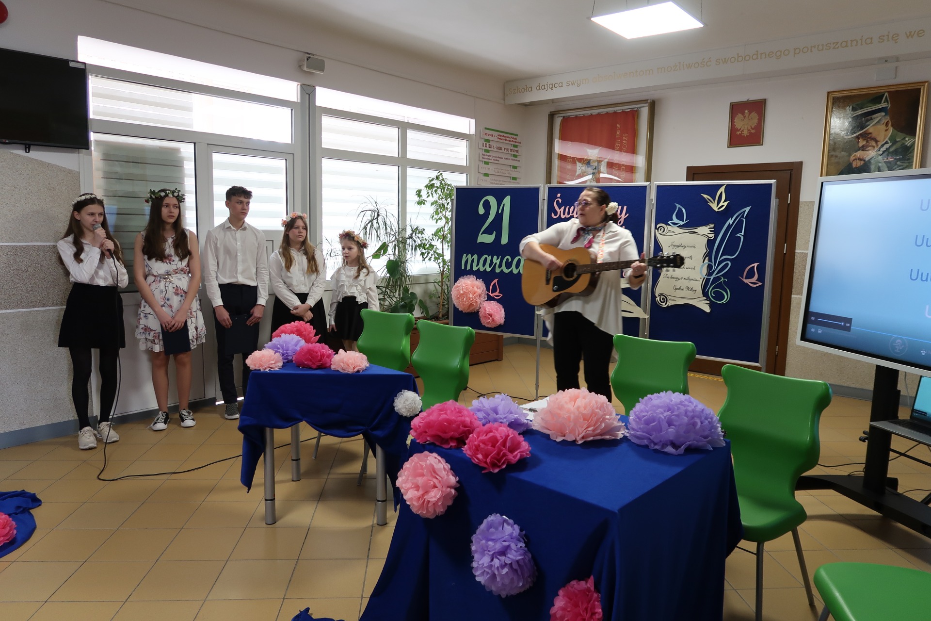 Uczniowie - lektorzy i soliści na niebieskim tle, ozdobionym kwiatami w odcieniach różu. Nauczycielka grająca na gitarze.