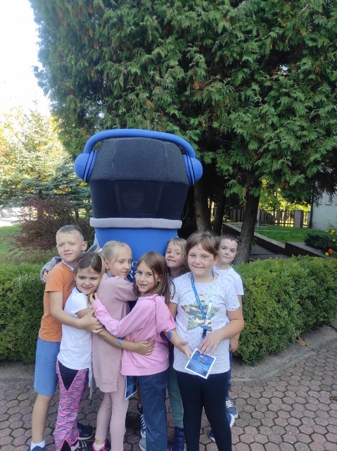 Siódemka dzieci stoi na zewnątrz, przytulają się do siebie i wielkiej maskotki mikrofonu
