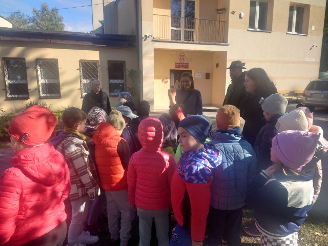 Uczniowie stoją w grupce przed budynkiem szkoły. Nauczycielki stoją przy uczniach. Leśnicy stoją przed dziećmi.