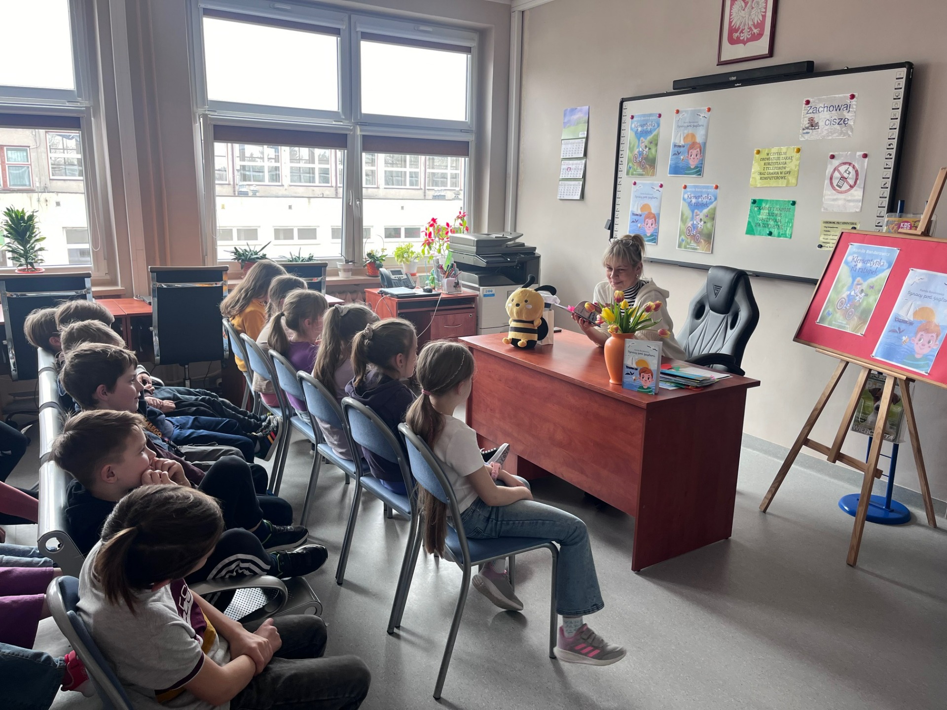Uczniowie siedzą w czytelni i słuchają autorki książki. 
