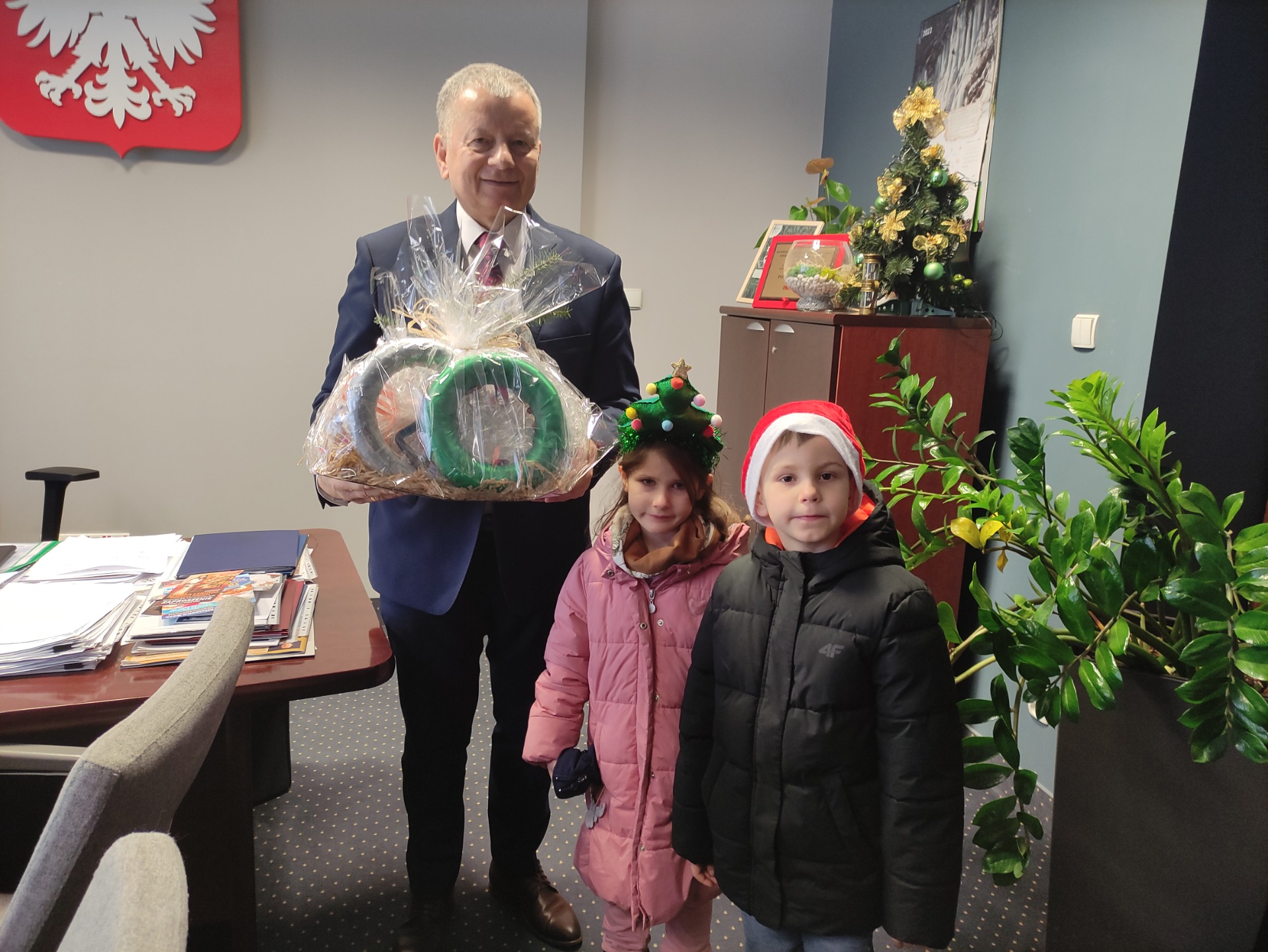 Zdjęcie przedstawia dwójkę dzieci w wieku 5 lat z Starostą 
Powiatu Mieleckiego Stanisławem Lonczakiem. Starosta trzyma w ręku otrzymany prezent od przedszkolaków ozdoby Bożonarodzeniowe wykonane podczas warsztatów z rodzicami.
