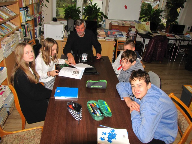 Przy stole siedzi nauczyciel, po jego prawej 3 uczennice, po lewej 3 uczniowie. Na stole leżą piórniki i książki. 