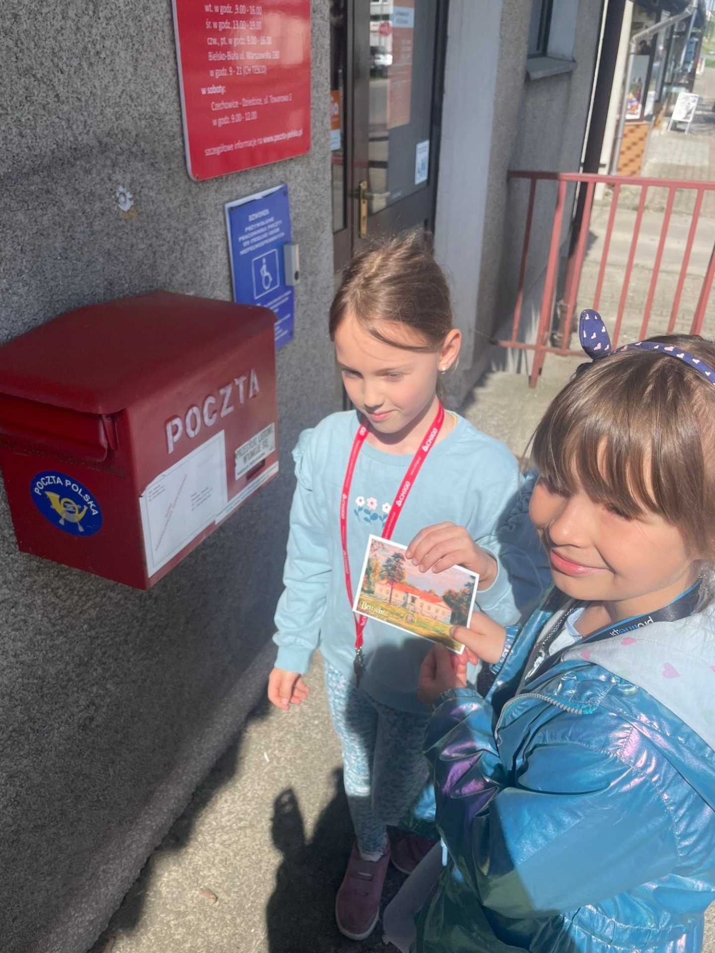 Dwie dziewczynki trzymają kartkę pocztową, w tle widać skrzynkę pocztową 
