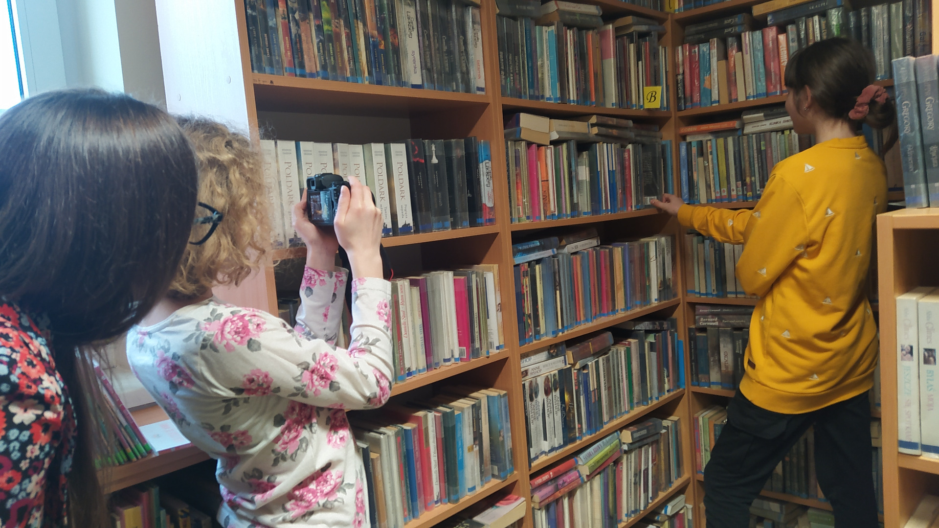 Uczennica wykonuje zdjęcie koleżance na tle półek z książkami w bibliotece