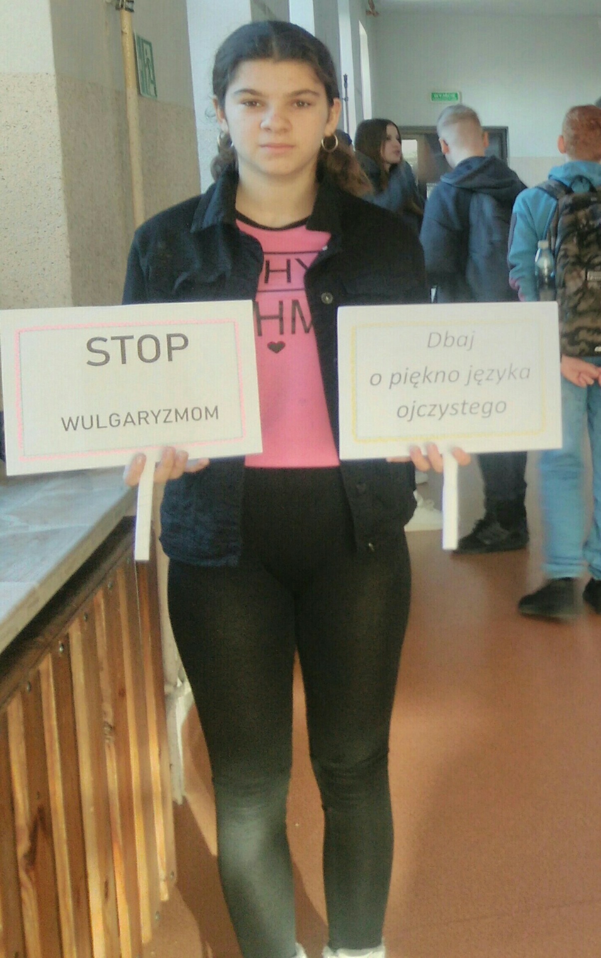 Uczennica klasy VII trzyma tabliczki z napisami: Stop wulgaryzmom i  Dbaj o piękno języka ojczystego.