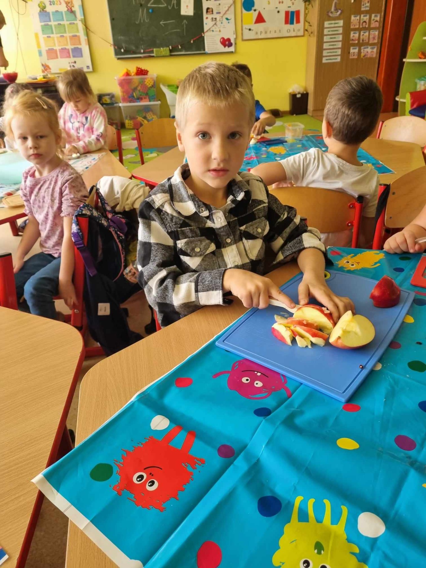 Przedszkolaki siedzą przy stolikach i kroją owoce na sałatkę