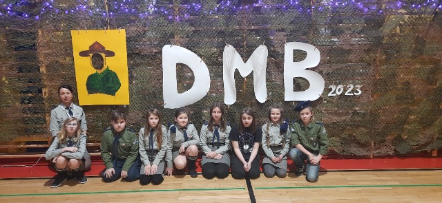 7 uczniów kuca na podłodze sali gimnastycznej. Za ich plecami znajduje się  napis DMB (Dzień Myśli Braterskiej) i plakat przedstawiający postać założycie skautingu Roberta Baden Powell'a. 