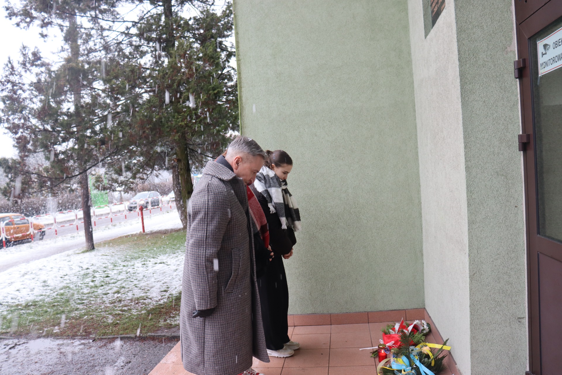 Mężczyzna oraz dwie uczennice stoją na dworze przed tablicą upamiętniającą Tragedię Górnośląską. Ich głowy są pochylone na znak hołdu. 