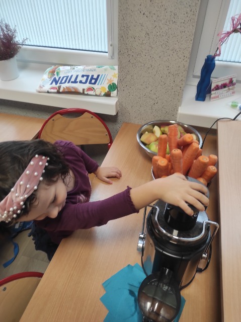 Dziewczynka robi sok w sokowirówkę, przechyla głowę i ogląda ten proces. Z boku widać marchewki i jabłka


